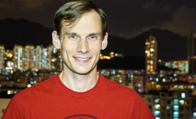 Male-presenting person in a red shirt standing in front of a nighttime cityscape.